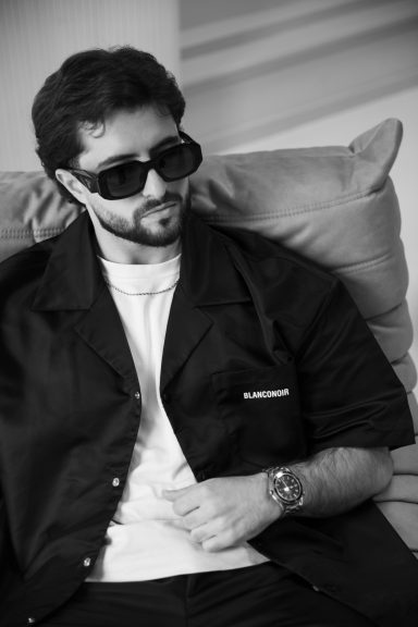 Man in zwarte jas en grote zonnebril, zittend op een bank in zwart-wit.