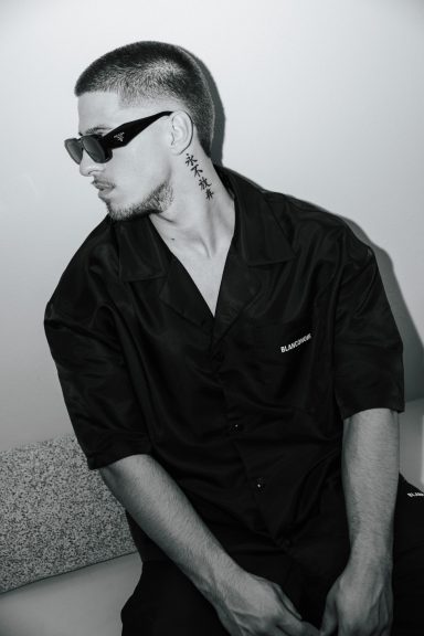 Man met kort haar en zonnebril, gekleed in een zwart shirt, zit op een bank.