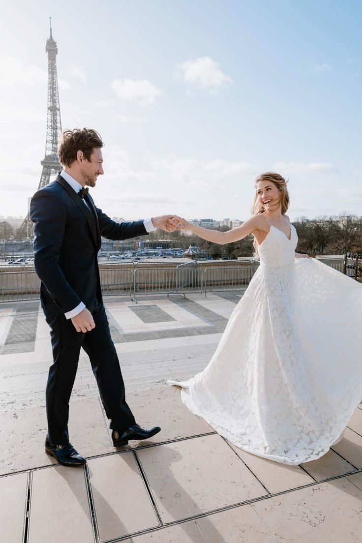Een stel in bruidskleding houdt elkaars handen vast met de Eiffeltoren op de achtergrond.