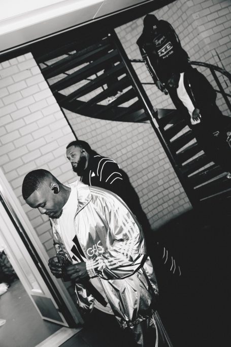 Drie mannen in sportkleding, staand bij een spiraaltrap in monochrome stijl.