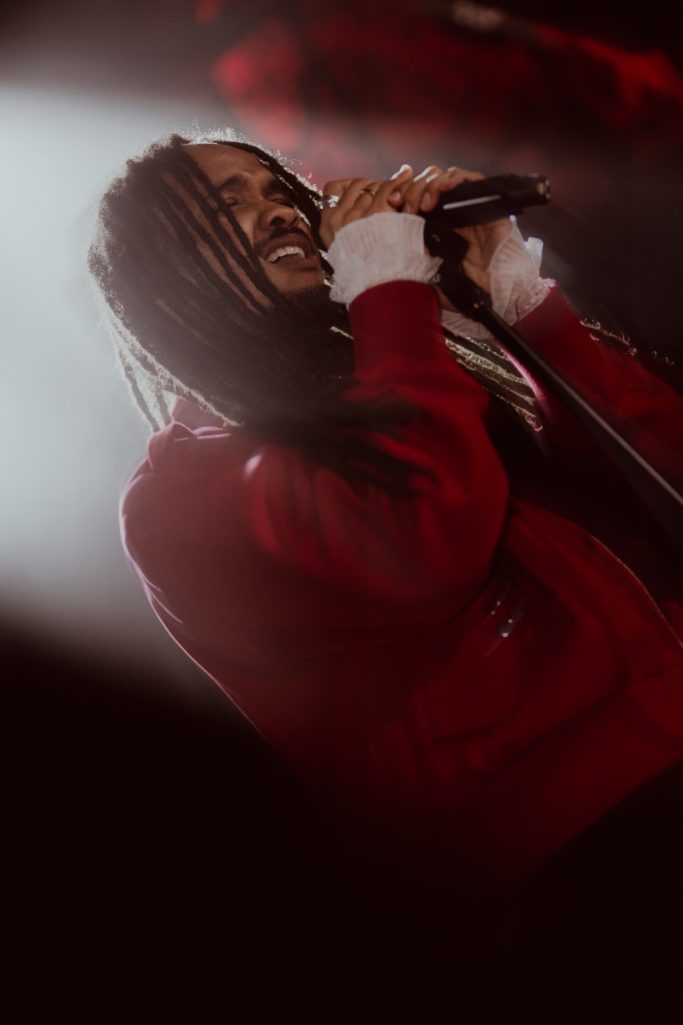 Jair Faria Bitterzoet Amsterdam Zanger met dreadlocks houdt een microfoon vast op een podium in zacht licht.