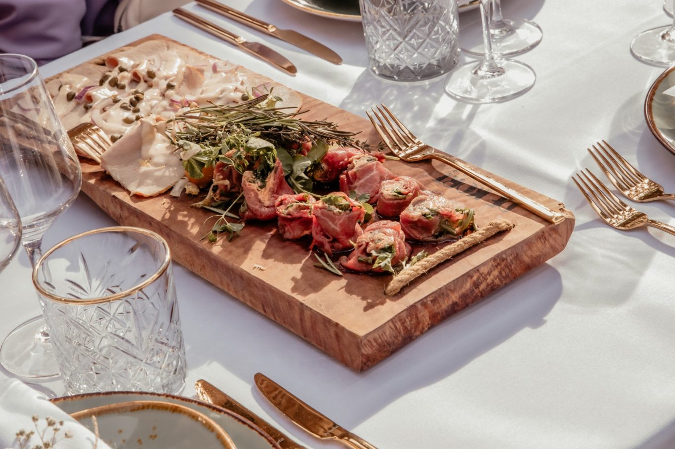 Houten plank met diverse hapjes, waaronder vlees, kruiden en kaas, op een elegante tafel.