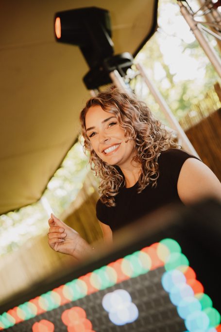 DJ H4zel - Geheime Liefde Festival Vrouw met krullend haar en een glimlach, staande achter een kleurrijke lichtinstallatie.