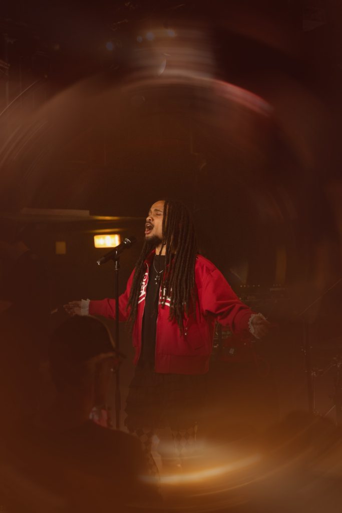 Jair Faria Bitterzoet Amsterdam Een persoon met lange dreadlocks in een rode jas staat staand met open armen.