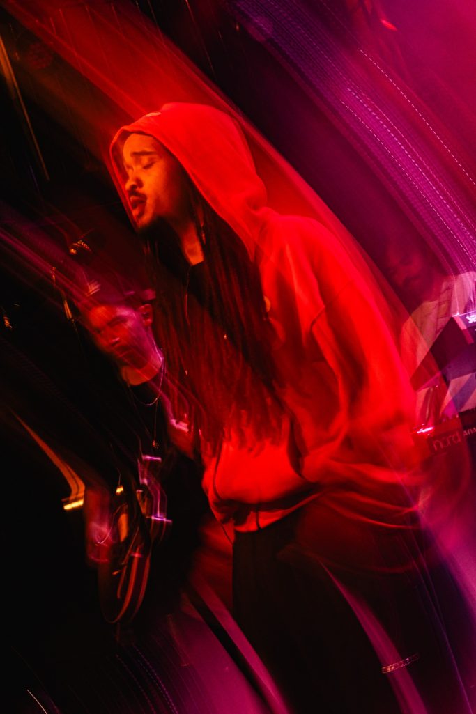 Jair Faria Bitterzoet Amsterdam Rehearsal Een persoon in een rode hoodie, met een ontspannen houding, op een podium met kleurrijke belichting.