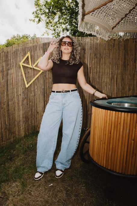 DJ H4zel - Geheime Liefde Festival Vrouw met krullend haar in een tuin, draagt een crop top en wijde jeans.