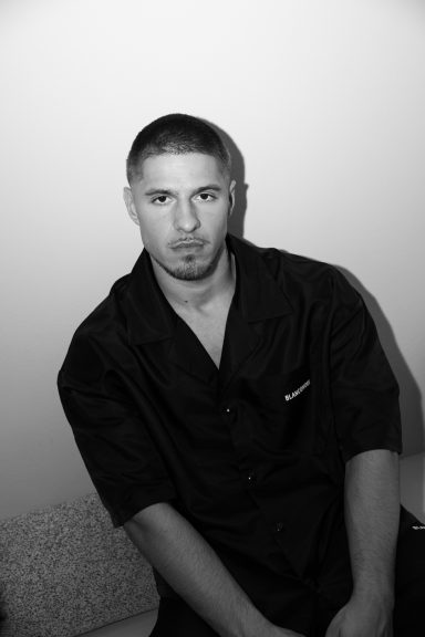 Man in een zwart overhemd, zittend met een neutrale uitstraling tegen een lichte achtergrond.