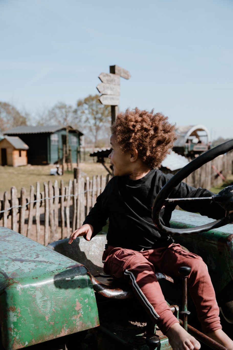 Een kind met krullend haar zit op een tractor, kijkt naar een boerderij in de verte.