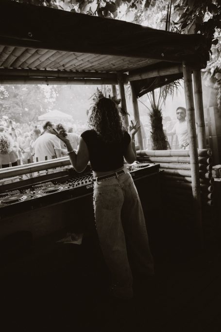 DJ H4zel - Geheime Liefde Festival Vrouw in zwart shirt en witte broek bij een kraam, met een natuurlijke omgeving.