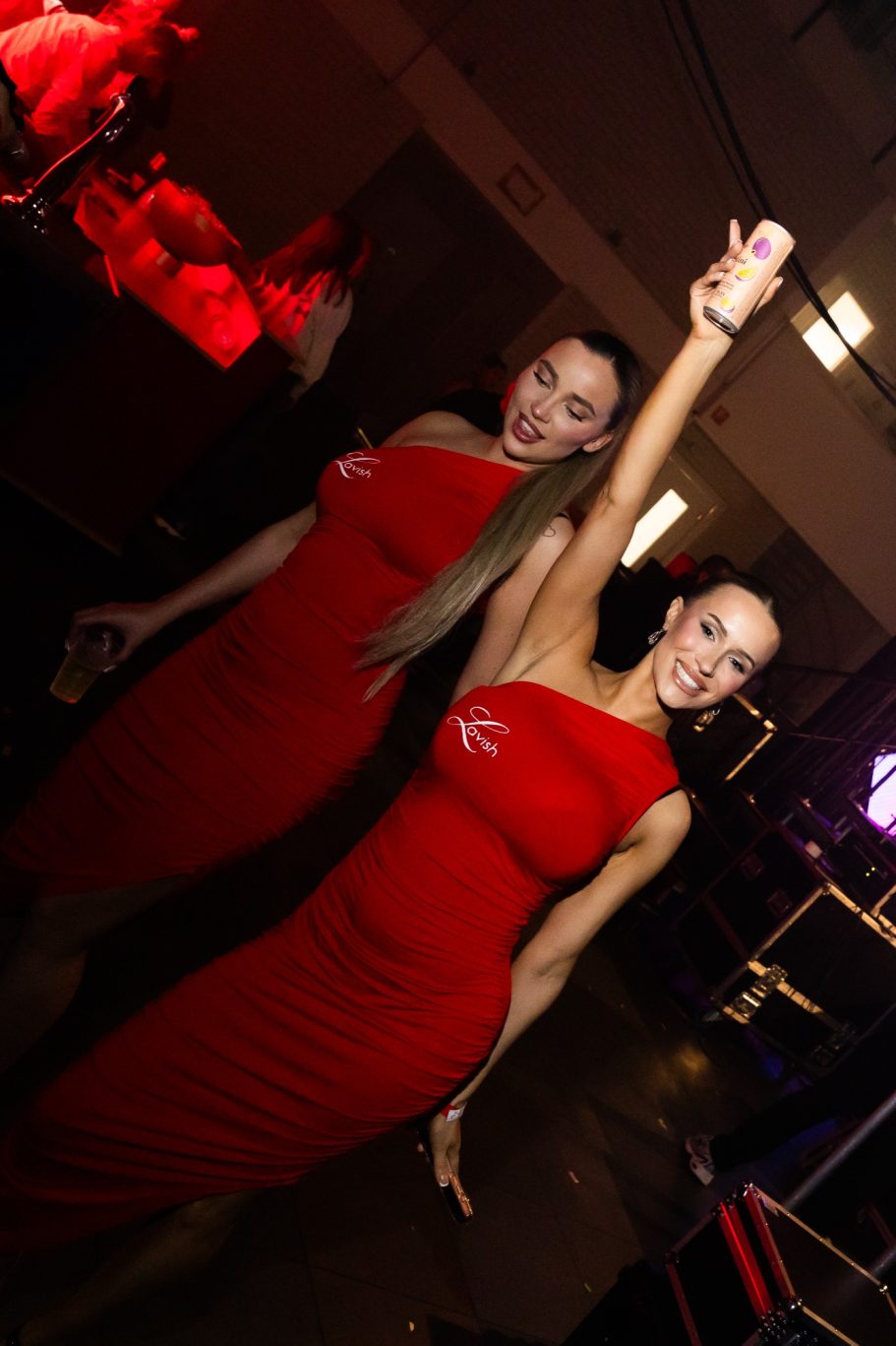 Twee vrouwen in rode jurken poseren met een hand omhoog, in een feestelijke omgeving.