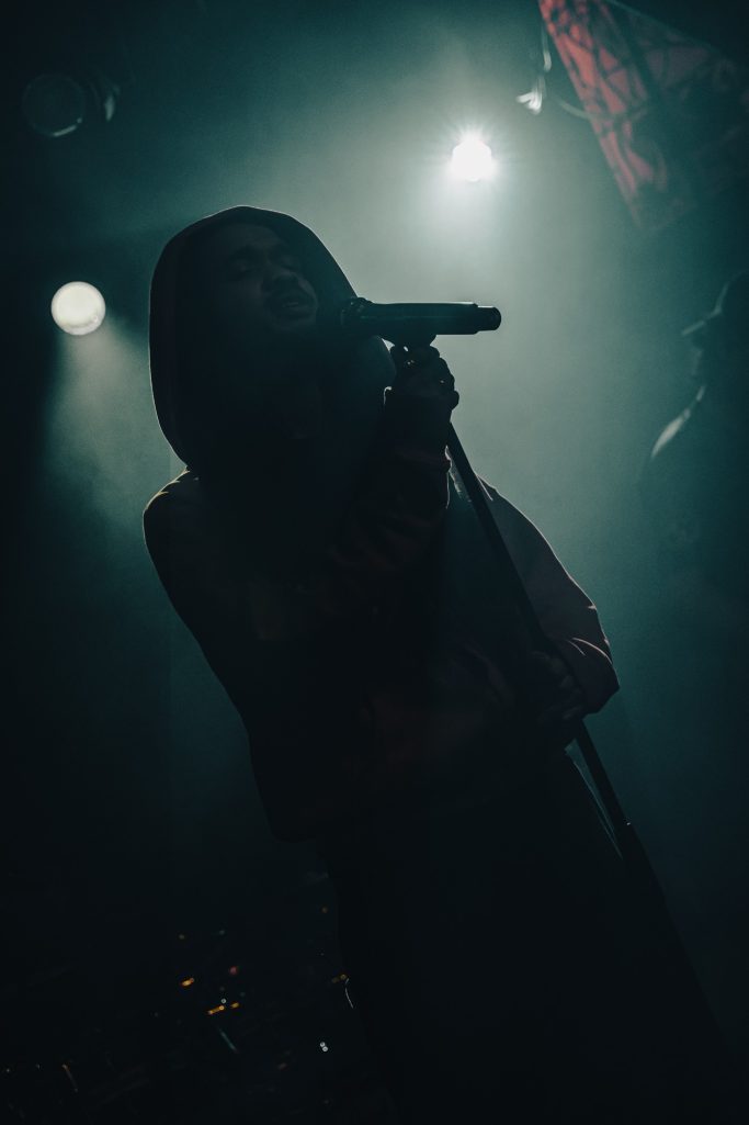 Jair Faria Bitterzoet Amsterdam Silhouet van een muzikant met microfoon, omgeven door sfeervolle verlichting.