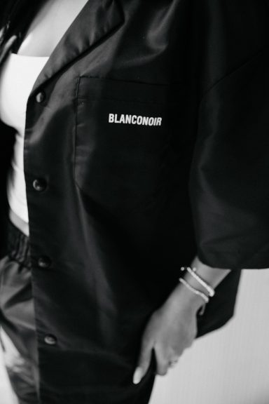 Zwart jack met opdruk "BLANCMANGE" en een hand met een armband.