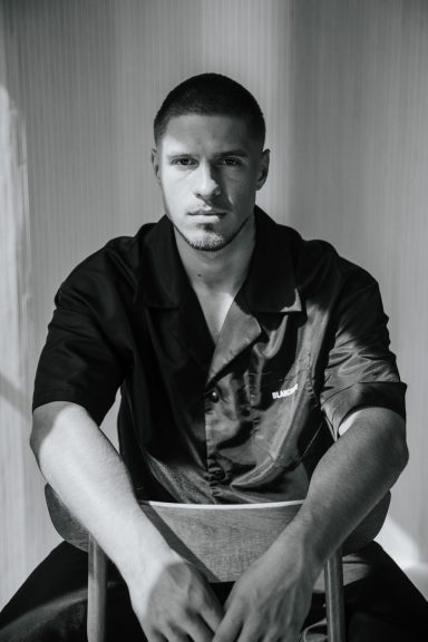 Een man kijkt serieus in de camera terwijl hij op een stoel zit.