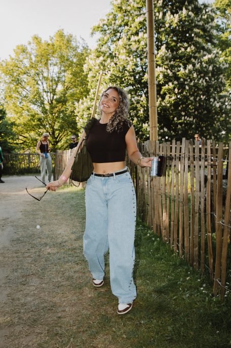 DJ H4zel - Geheime Liefde Festival Jonge vrouw met krullend haar, draagt een zwart crop-top en lichte spijkerbroek, glimlachend.