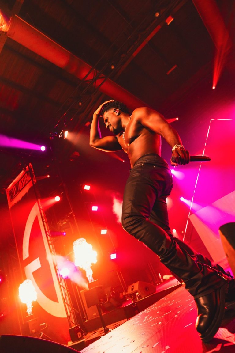 Zanger op podium met dramatische belichting en vlammen, in een energieke pose.