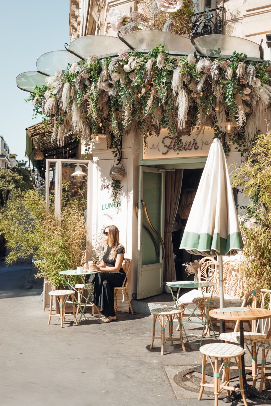 Een vrouw zit buiten een café met planten en een parasol.