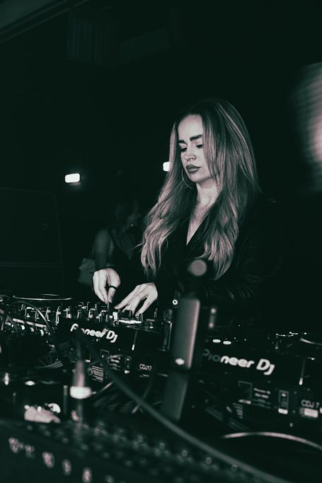 Vrouwelijke DJ met lang haar, geconcentreerd achter de draaitafels in een donkere setting.