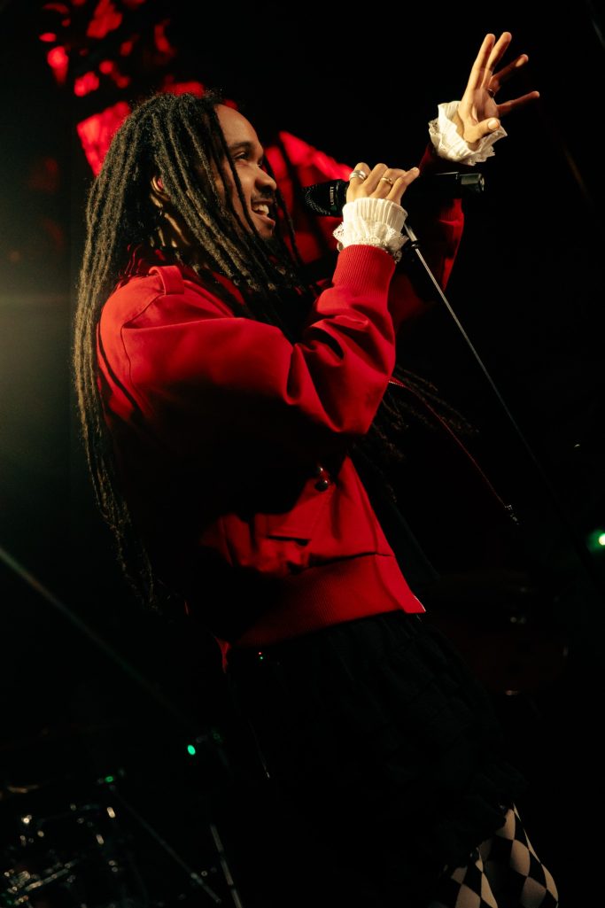 Jair Faria Bitterzoet Amsterdam Zanger met dreadlocks, in een rode trui, die optreedt met een microfoon.