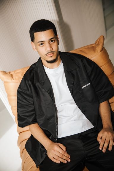 Man in zwart shirt en witte T-shirt, zittend op een kussen met een neutrale achtergrond.