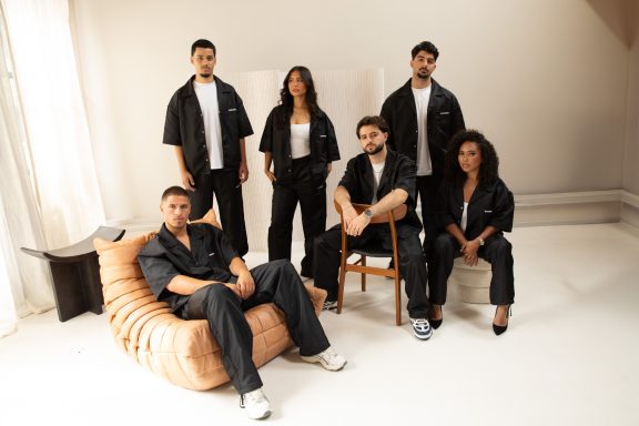 Zes personen in stijlvolle zwarte outfits zitten en staan in een lichte ruimte.
