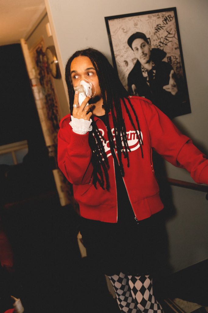 Jair Faria Bitterzoet Amsterdam BTS Een persoon in een rode hoodie en zwart-witte broek, staat met een drankje in de hand.