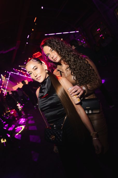 Twee vrouwen poseren met glamoureuze outfits op een feestelijke locatie met kleurrijke verlichting.