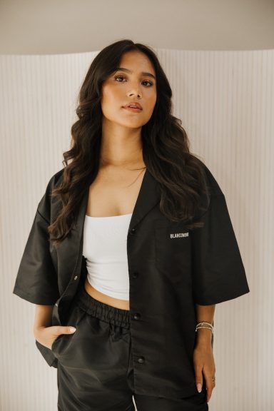 Vrouw met lange, golvende haren in een zwart overhemd en broek, staand voor een lichte achtergrond.