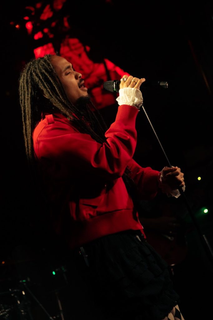 Jair Faria Bitterzoet Amsterdam Man met dreadlocks in een rode trui, zingt in een microfoon op een podium.
