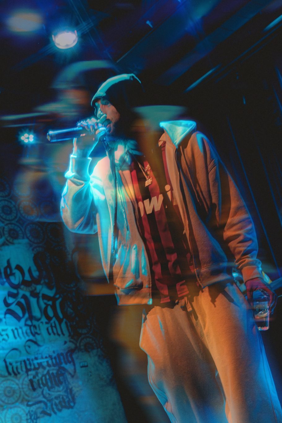 Rapperformer met microfoon op een podium, omgeven door kleurrijke lichten.