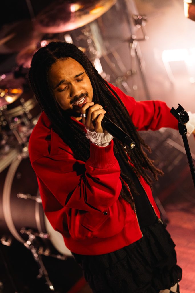 Jair Faria Bitterzoet Amsterdam Zanger met lange dreadlocks in een rode jas, optredend op een podium met muzikanten.
