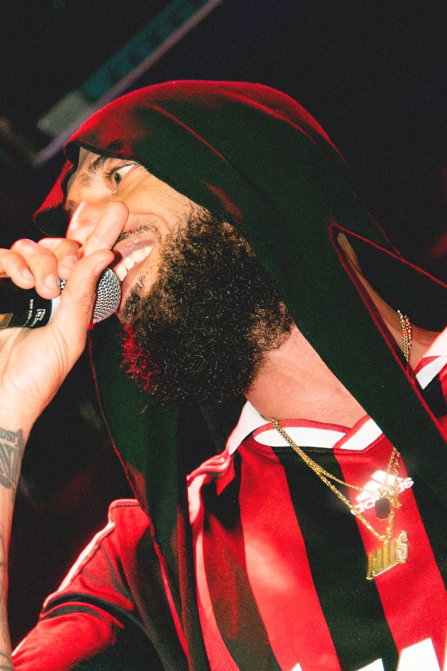 Man in een zwart-rood shirt zingt in een microfoon met een capuchon over zijn hoofd.