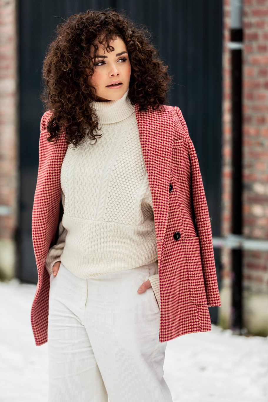 Vrouw met krullend haar in een roze jas en lichte kleding, staand op sneeuw.