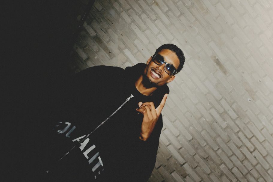 Jonge man met zonnebril, glimlachend en een handgebaar in een donkere omgeving.