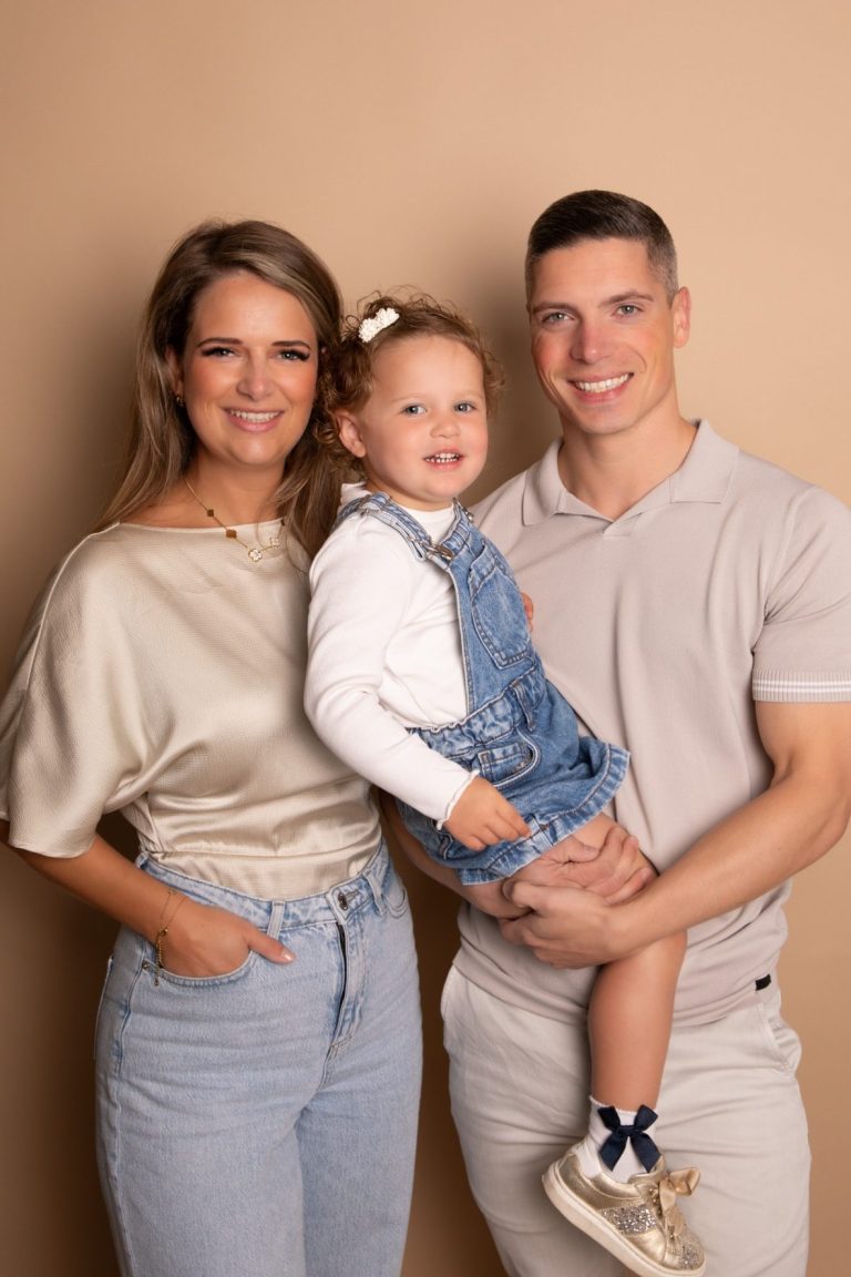 Een gezin met een meisje in een spijkerbroek, staand voor een beige achtergrond.