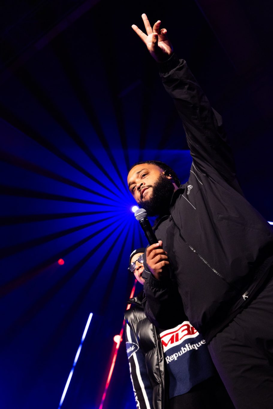 Persoon op het podium met microfoon, verlicht door stralen van blauw licht.