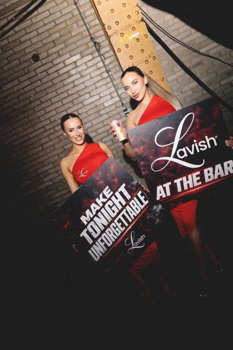 Twee vrouwen in rode jurken houden borden vast voor een evenement in een bar.