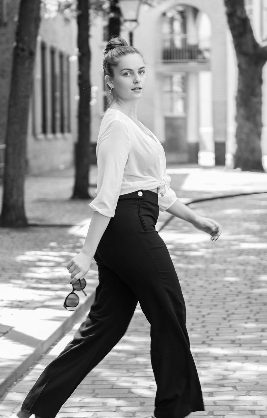 Een vrouw in een witte blouse en zwarte broek wandelt ontspannen op een straat.