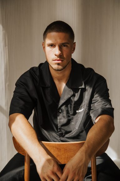 Man in zwart kleding zit op een houten stoel, met duidelijke licht- en schaduwspel.