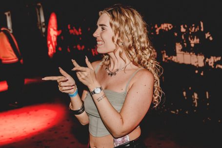 Jonge vrouw met krullend haar, wijst en lacht in een sfeervolle, verlichte ruimte.