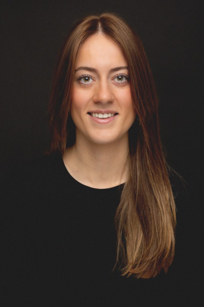 Portret van een vrouw met lang bruin haar, lachend tegen een donkere achtergrond.