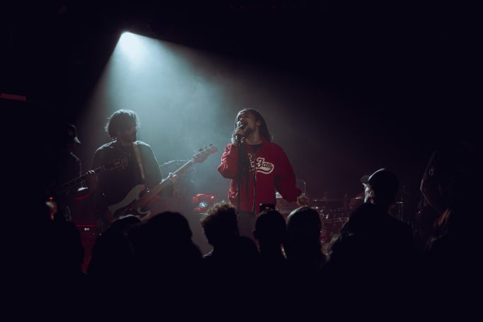 Jair Faria Bitterzoet Amsterdam Zanger met een rode trui performt op podium, met band en publiek in de spotlight.