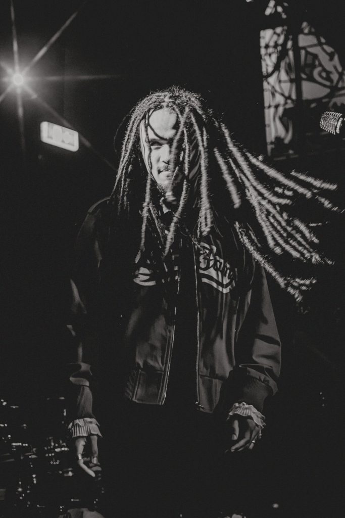 Jair Faria Bitterzoet Amsterdam Man met dreadlocks in een donkere setting, draagt een zwart jack.