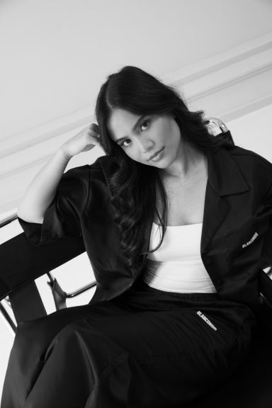 Jonge vrouw in een zwart outfit, zittend op een stoel, met een zachte uitstraling.