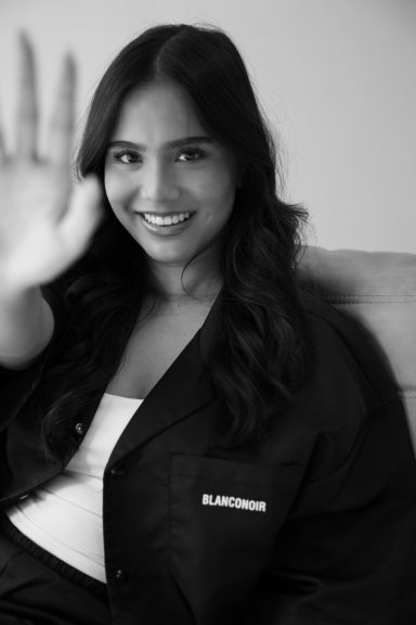 Vrouw met een lach en opgestoken hand, gekleed in een zwart jasje, tegen een neutrale achtergrond.