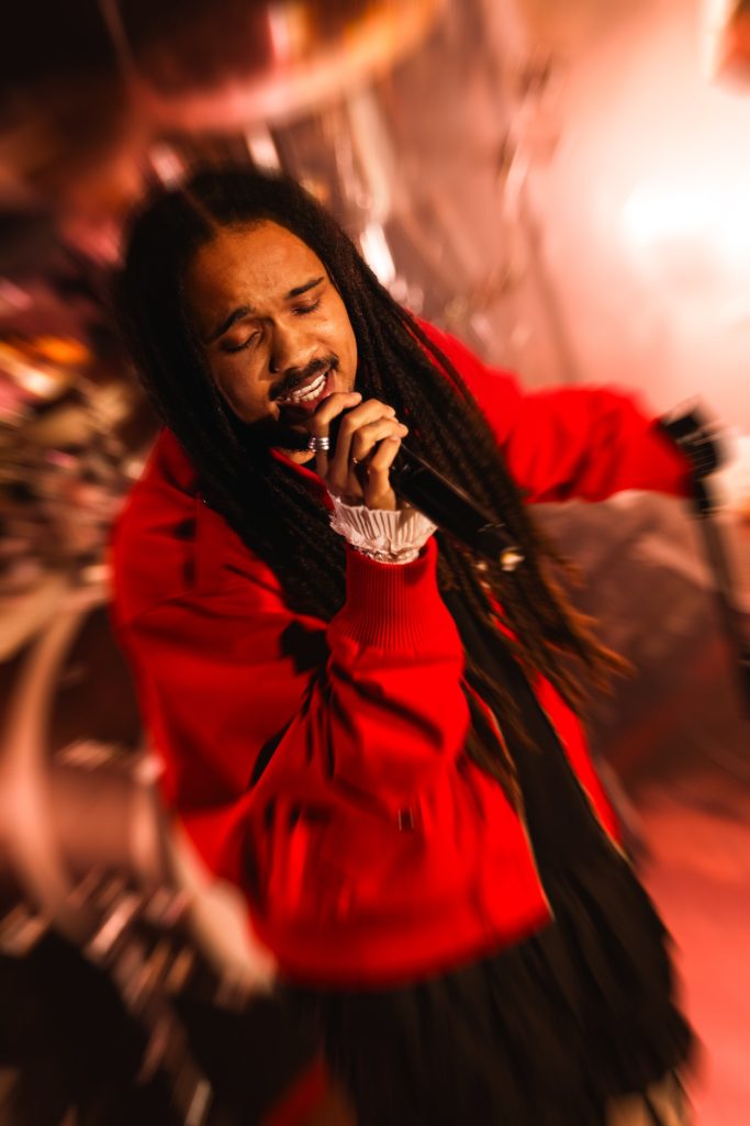 Jair Faria Bitterzoet Amsterdam Zanger met dreadlocks in een rode jas, zingt op het podium met microfoon.