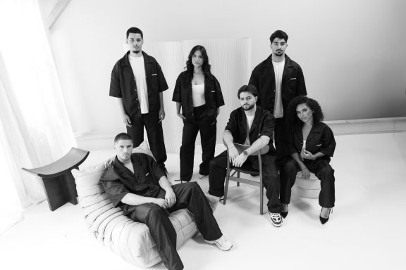 Zes mensen in zwarte kleding zitten en staan in een lichte, moderne ruimte.