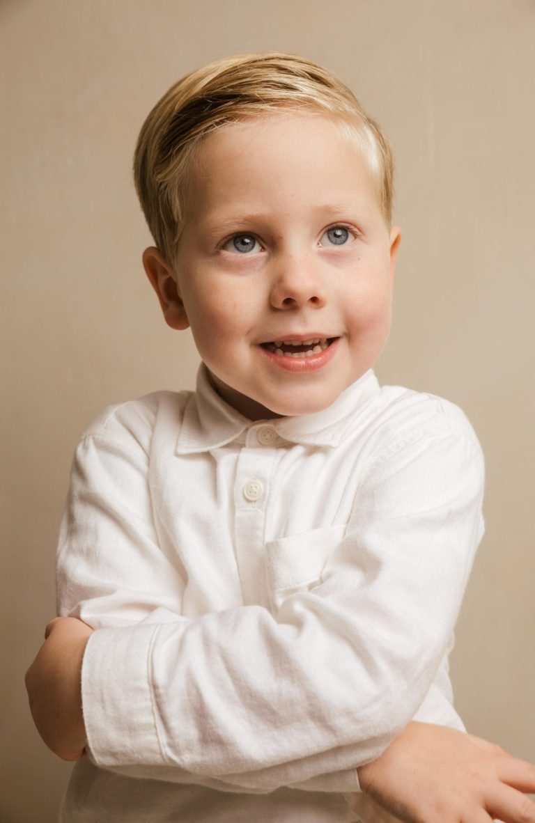 Jonge jongen met een blije glimlach en gekruiste armen, gekleed in een witte blouse.