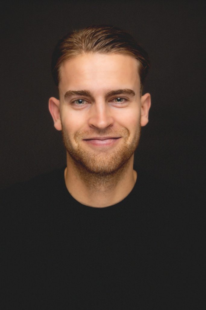 Man met een vriendelijke glimlach, kort haar en een zwart T-shirt op een donkere achtergrond.