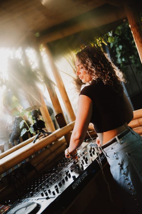 DJ H4zel - Geheime Liefde Festival Vrouw met krullend haar achter een DJ-console in een groene, zonnige omgeving.