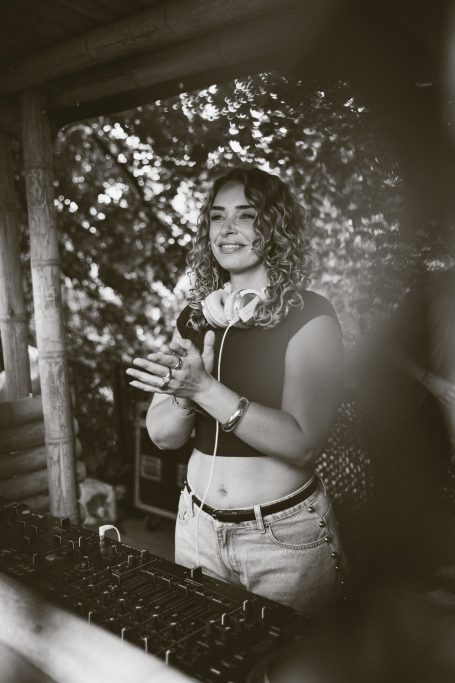 DJ H4zel - Geheime Liefde Festival Jonge vrouw met krullend haar, glimlachend en met handen in de lucht, omgeven door groen.